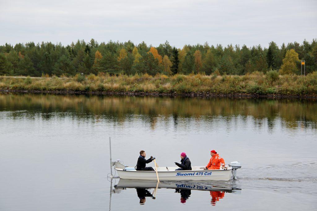 Syksyinen jokimaisema, veneessä kolme henkilöä tekemässä tutkimusta. 