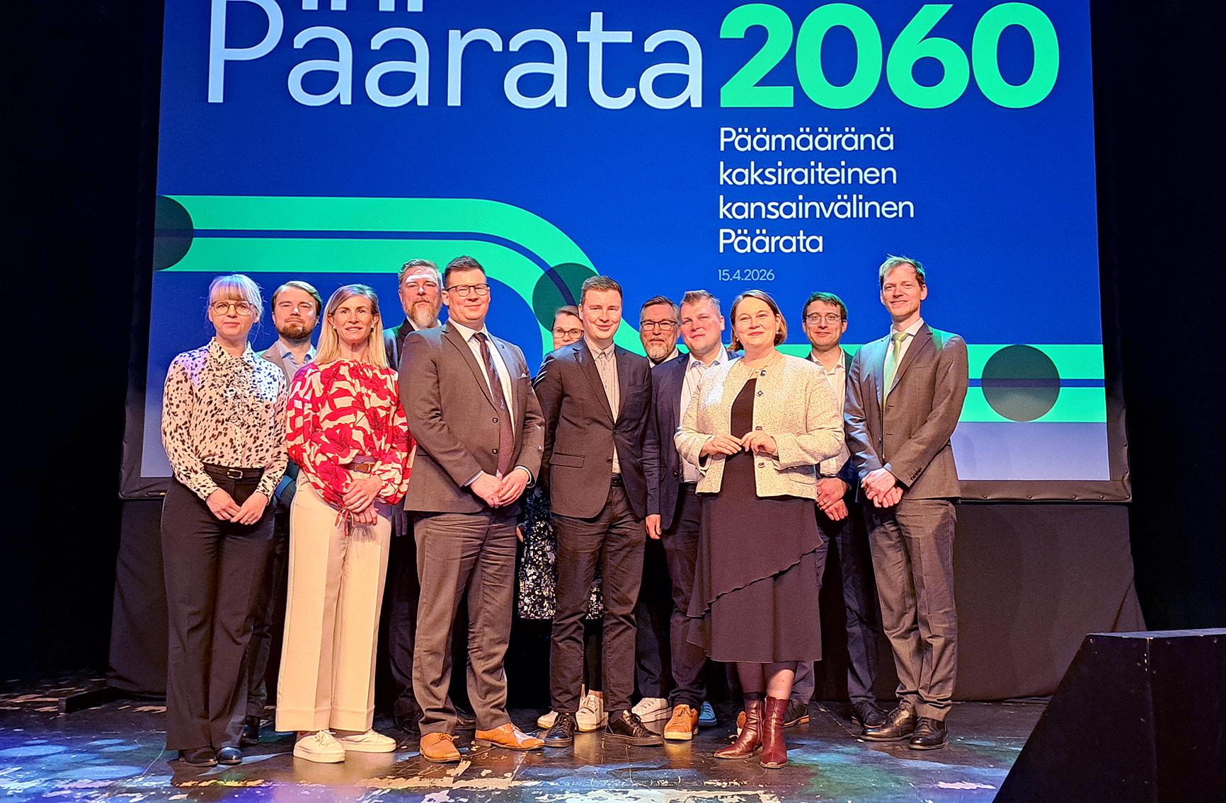 Sinivihreän taustan edessä seisoo joukko ihmisiä ryhmäkuvassa. Takana on isolla teksti Päärata2060 - päämääränä kaksiraiteinen kansainvälinen päärata.