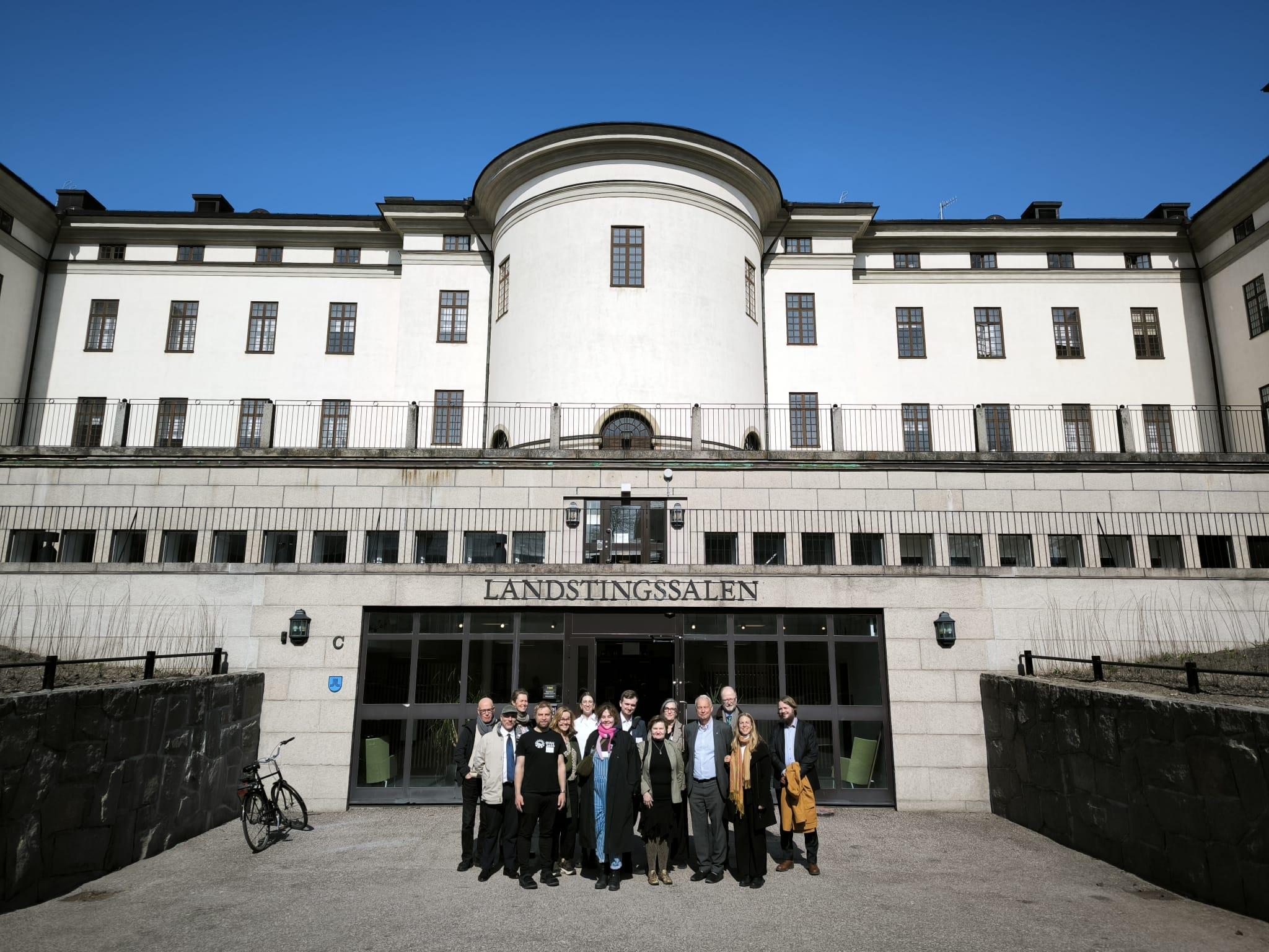 Ryhmä ihmisiä seisoo valkoisen vanhan kivitalon edustalla. Talo on Tukholman läänintalo.