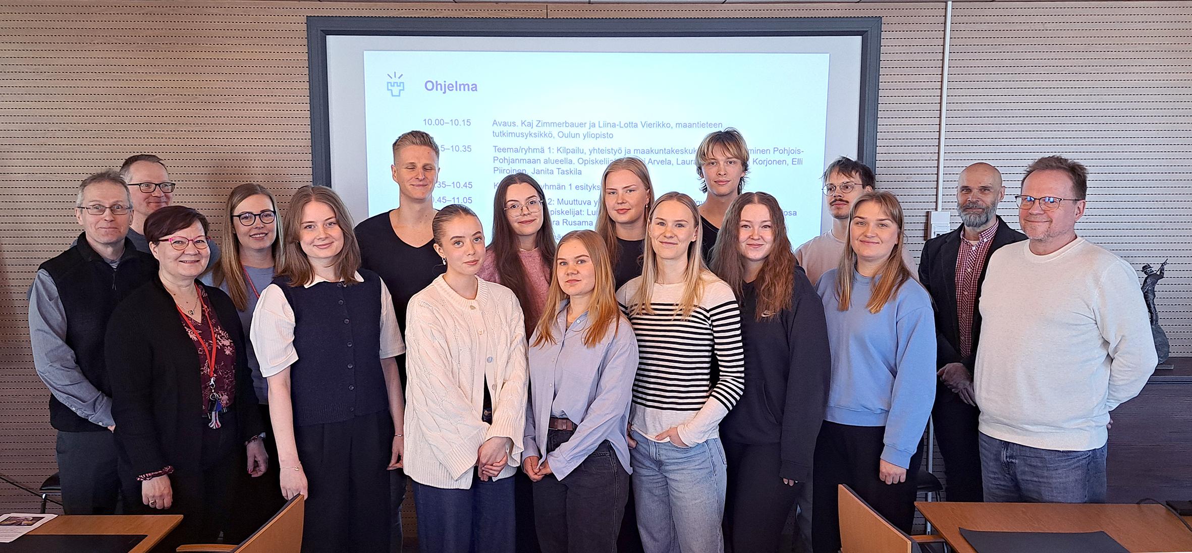 Iloinen ryhmä ihmisiä seisoo rykelmässä. Taustalla vaalean ruskea seinä sekä powerpoint -esitys.