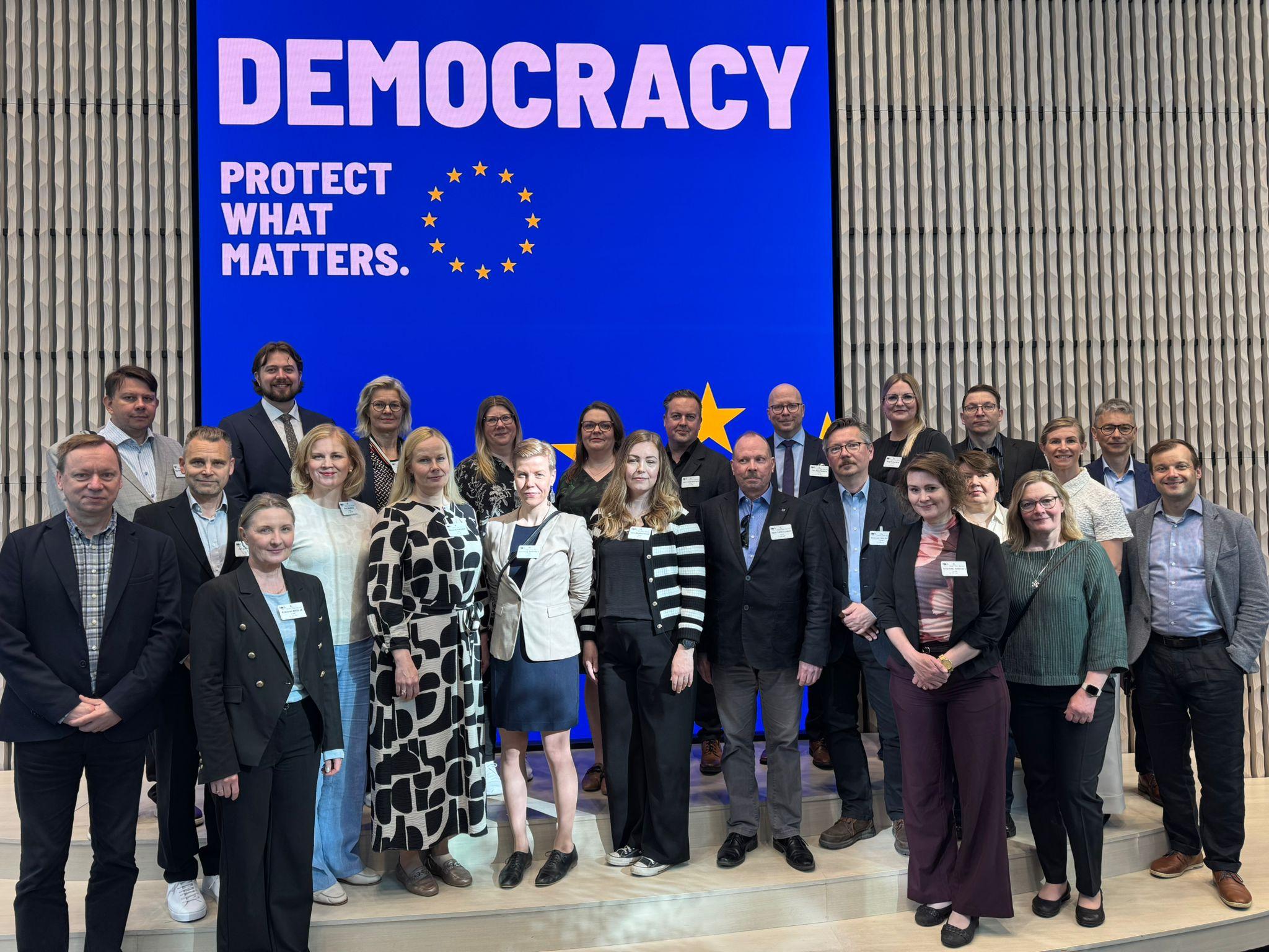 Ryhmä ihmisiä ryhmäkuvassa. Henkilöitä seisoo kolmessa rivissä. Taustalla rimaseinä, sekä sininen näyttö, jossa EU:n tähdet ja teksti Democracy. Protect what matters.