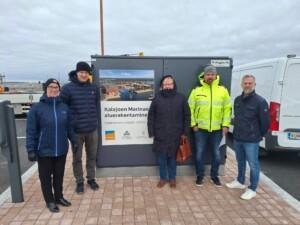 Viisi ihmistä seisoo sähköpääkeskuksen edessä. Taustalla on sähköpääkeskukseen kiinnitetty kyltti, jossa lukee Kalajoen Marinan aluerakentaminen.