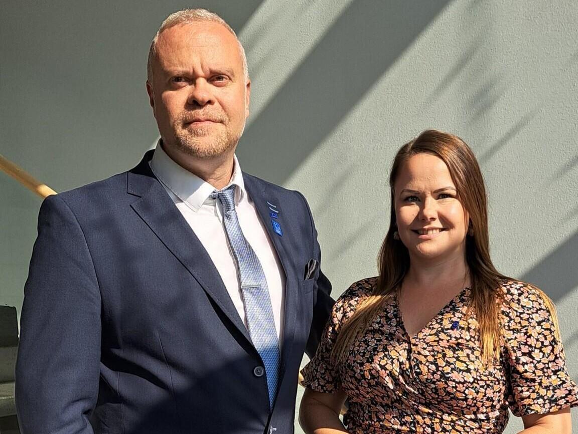 Ruskeahiuksinen nainen seisoo kesämekossa kädet yhteen liitettynä vierellään tummansiniseen pukuun pukeutunut mies.